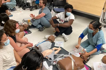 Students in a simulation center sit on the floor. A facilitator is demonstrating how to give CPR.