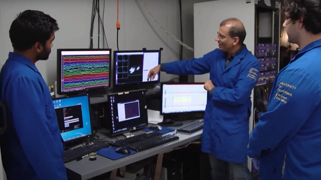 A group of three men in a lab stand around monitors with data onscreen, the older of the three points to the screen in explanation.