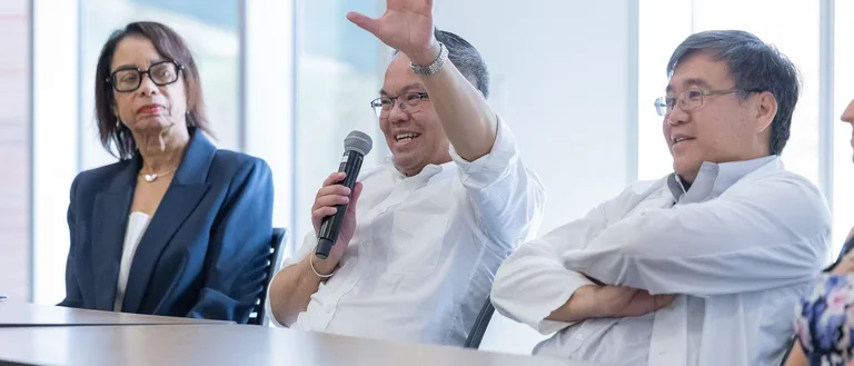Several individuals gathered around a table, one person actively raising their hand to contribute to the discussion