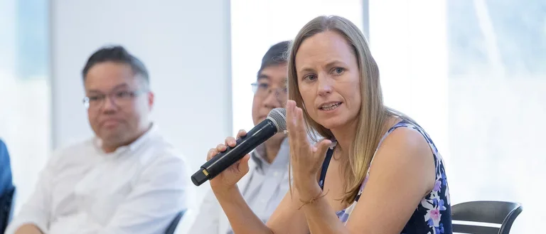 A woman speaks into a microphone while seated at a table, engaging her audience with confidence.