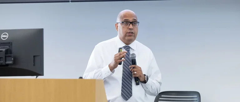 A man wearing a white shirt speaks to a gathering of people, sharing ideas and insights.