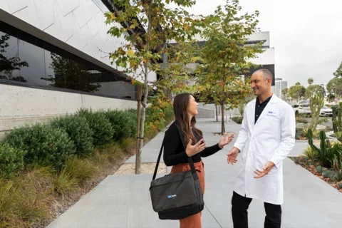 A man in a lab coat and a woman with a black bag chat on a tree-lined walkway outside a modern building, with city buildings in the background.