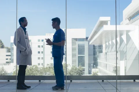 A UCLA Health resident and physician confer in front of a large window looking out over south campus at UCLA