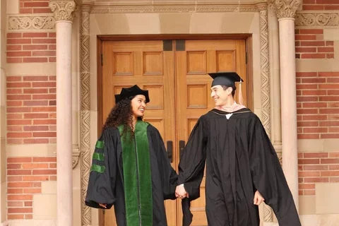 UCLA residents and med-school alumni Makana Williams and Rigoberto Perez Hernandez smile at each other wearing graduation robes in front of an academic building. 