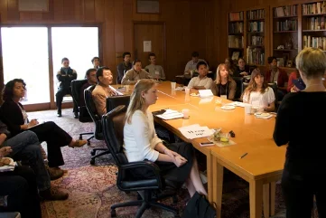 Group of people sitting around a table inside a room with a person standing at the front