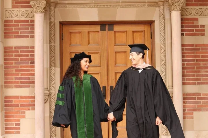 UCLA residents and med-school alumni Makana Williams and Rigoberto Perez Hernandez smile at each other wearing graduation robes in front of an academic building. 