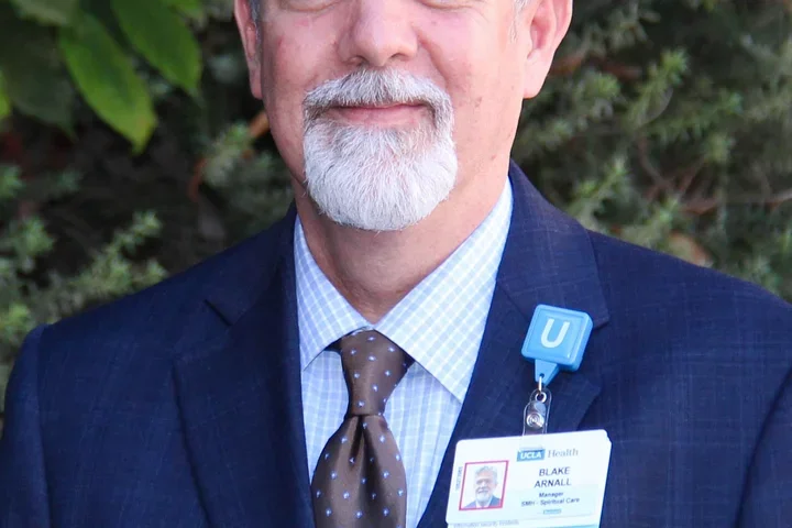 Gray-haired bearded man in a blue suit with an ID badge, standing outdoors.