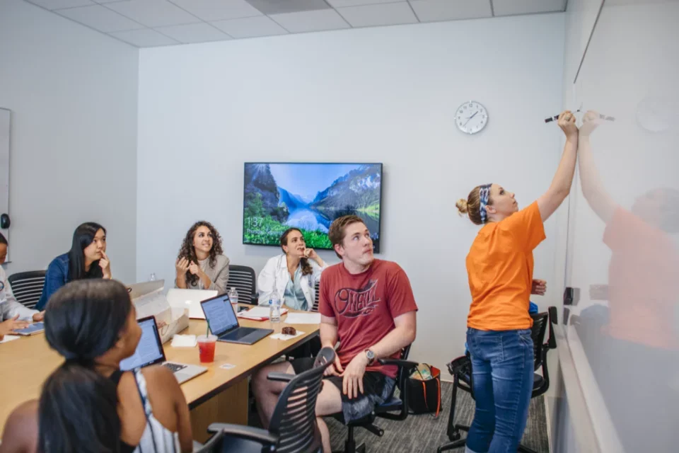 A group of pre med students in a teams based learning classroom. One writes on a whiteboard while the others watch.