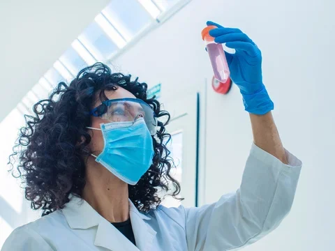 Woman in surgical mask and lab goggles holding up lab tube
