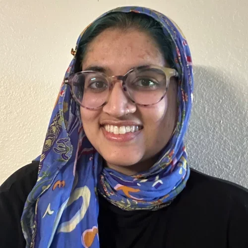 girl with glasses in a blue headscarf and black t-shirt standing in front of a white wall