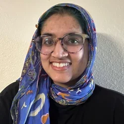 girl with glasses in a blue headscarf and black t-shirt standing in front of a white wall