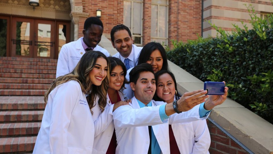 A group of medical students wearing white coats pose for a selfie after the white coat ceremony.