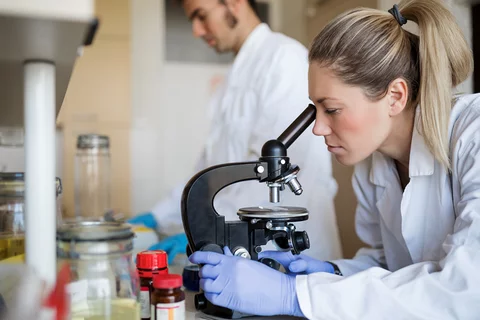 A woman looking into a microscope with a lab mate behind her