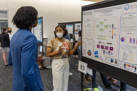 A medical student reviews her work at the Senior Scholarship Day. She's standing in front of her research poster and explaining to an onlooker.