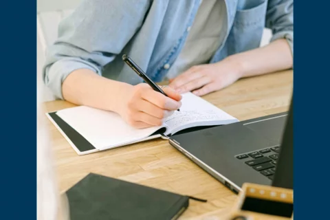 Man writing on a piece of paper in front of a laptop with stationary around him.
