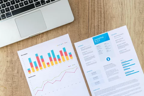 a graph and a resume on a brown wooden table with a laptop next to them in the upper left corner