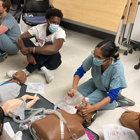Students in a simulation center sit on the floor. A facilitator is demonstrating how to give CPR.